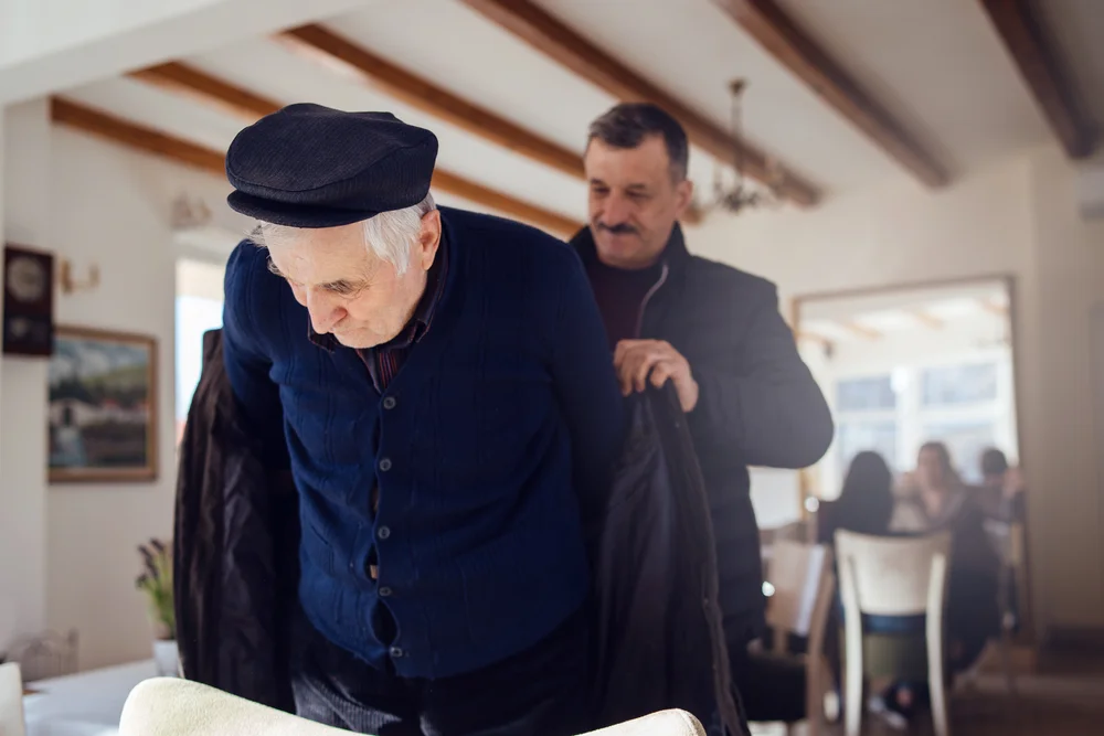 Senior man 80 years old caucasian pensioner and his son 60 years old man helping him to get dressed holding the coat at the restaurant in sunny day nursery retirement home
