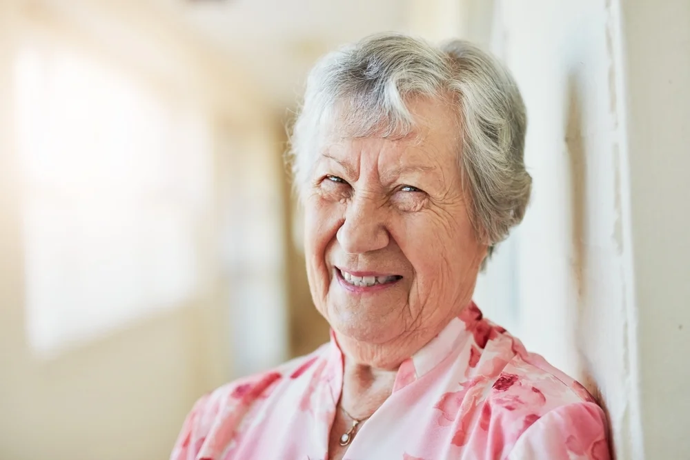Elderly woman, face and smile in retirement home by corridor wall for peace, health and positive mood. Senior person, portrait and happy in nursing house in hallway for wellbeing, calm and gratitude