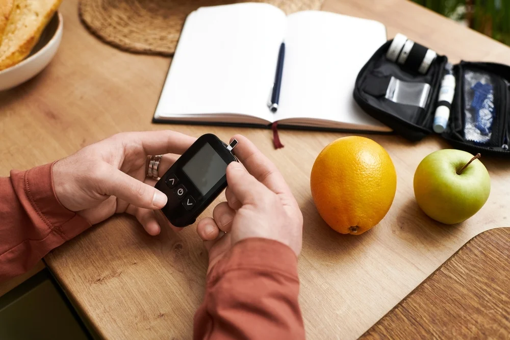 A young man checks his blood sugar levels at home surrounded by healthy snacks and planning tools.