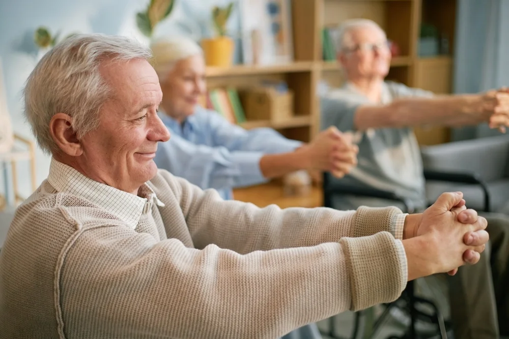 Elderly people in retirement home performing stretching exercises while seated, focusing on health and well-being amid light-filled indoor setting with plants and bookshelves