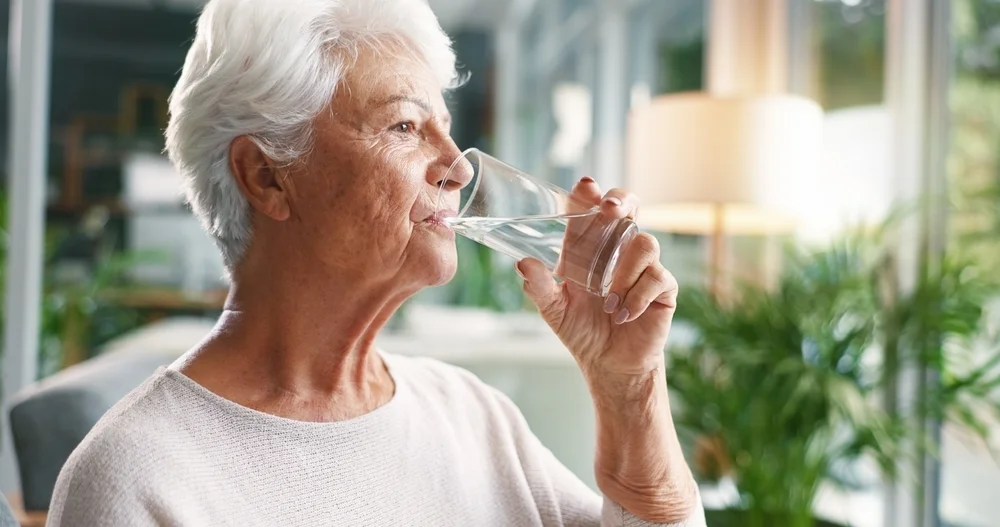 Home, drinking water and old woman in lounge, wellness and hydration with morning routine. Nutrition, pensioner and elderly person with liquid, thirsty and aqua with health in retirement and detox