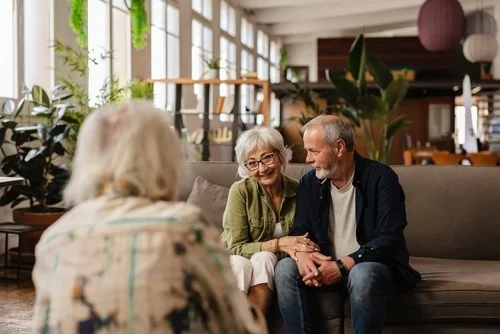 Happy senior couple holding hands and engaging in conversation with an elderly friend while sitting comfortably on the sofa in their cozy living room. Enjoying moments of togetherness and connection