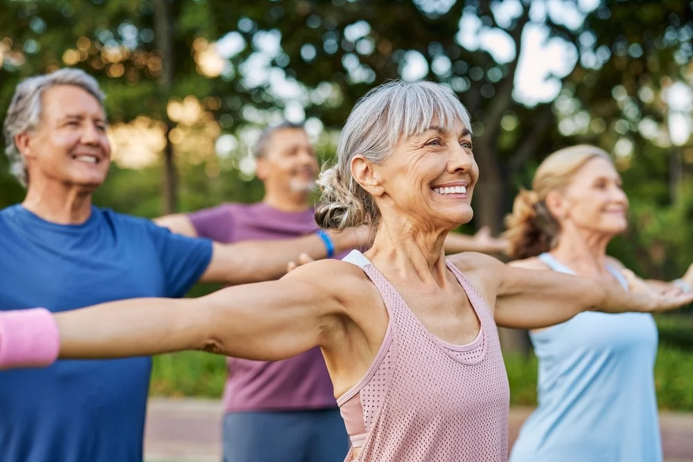 Happy senior group doing yoga together in the park, gentle gymnastics. Mature men and women stretching arms in outdoor fitness class. Sporty elderly friends doing a arm warm up workout before training