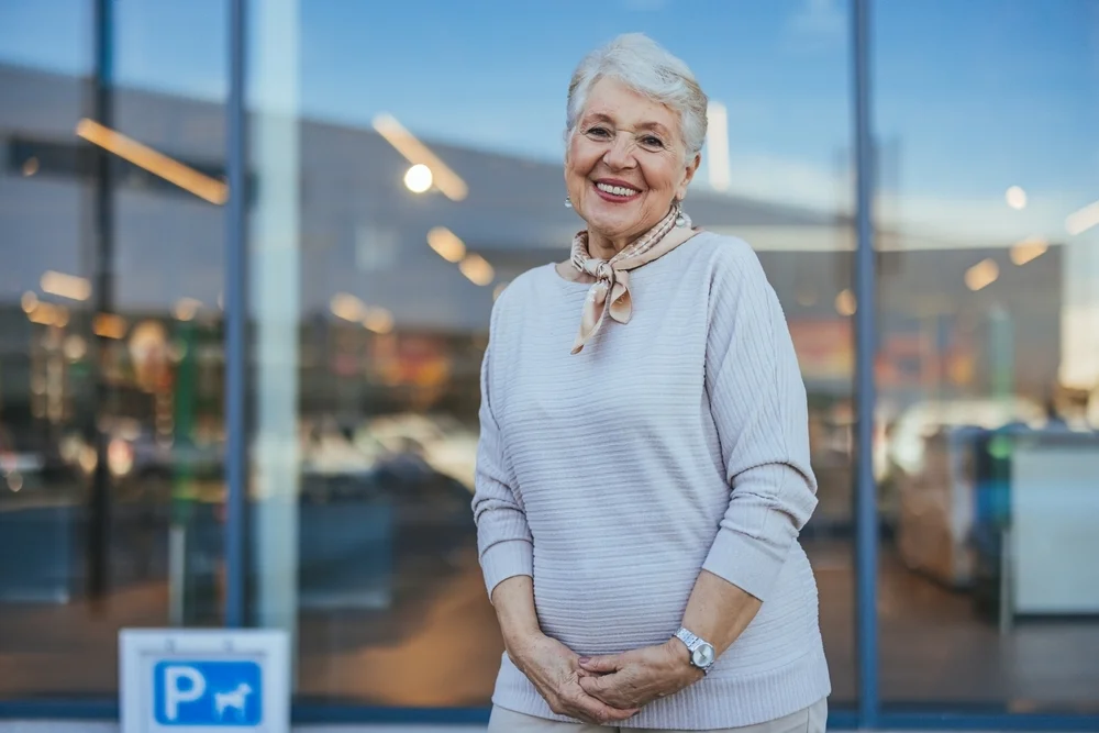 An elderly woman with a joyful expression stands confidently outside a modern glass building. Her welcoming smile radiates warmth and positivity, embodying happiness and vitality in her senior years.