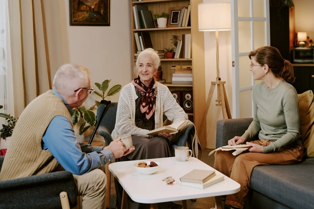 Senior Caucasian woman sitting between middle aged Caucasian man and middle aged Caucasian woman, holding book and smiling while engaging in conversation in living room setting