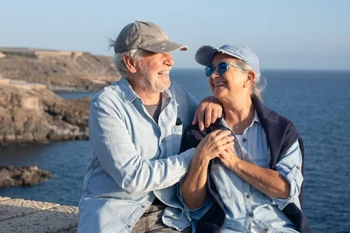 Happy senior couple hugging and smiling while sitting on a stone wall by the sea. Enjoying retirement, love, and companionship during a sunny coastal vacation