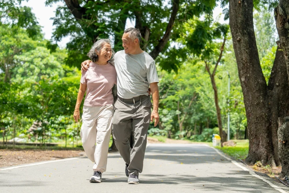 Asian senior man put his arm around senior woman and walk along the road in public park and they look each other with happiness during exercise and relax together.