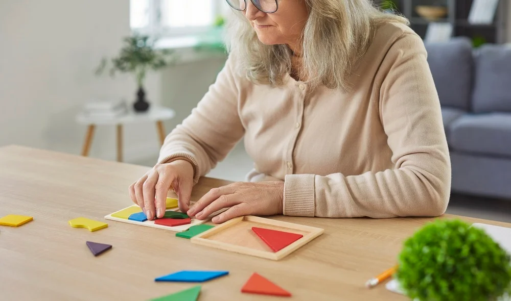 Senior old hobby, older woman sitting at table, solving tangram puzzle, arranging geometric shapes, focused and calm, engaging in cognitive activity, mentally alert, physically fit, therapeutic class